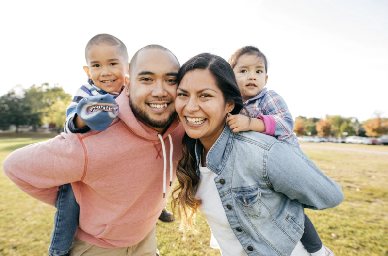 “A happy young family of four smiles at the camera while playing outside in a park. The parents carry their two children on their backs, symbolizing joy, togetherness, and a bright future.”
