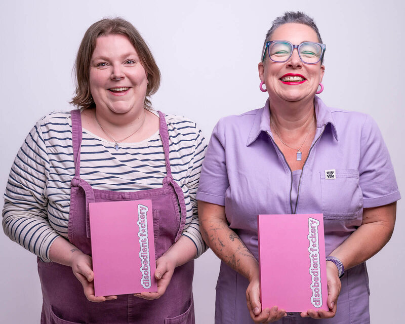 Two people grin at the camera against a pale studio backdrop, each holding a bright pink notebook labelled “disobedient f*ckery.” Lucy on the left wears a striped top with mauve dungarees; Pippa on the right wears lilac collared dungarees, blue rimmed glasses, pink hoop earrings, and has tattooed forearms.
