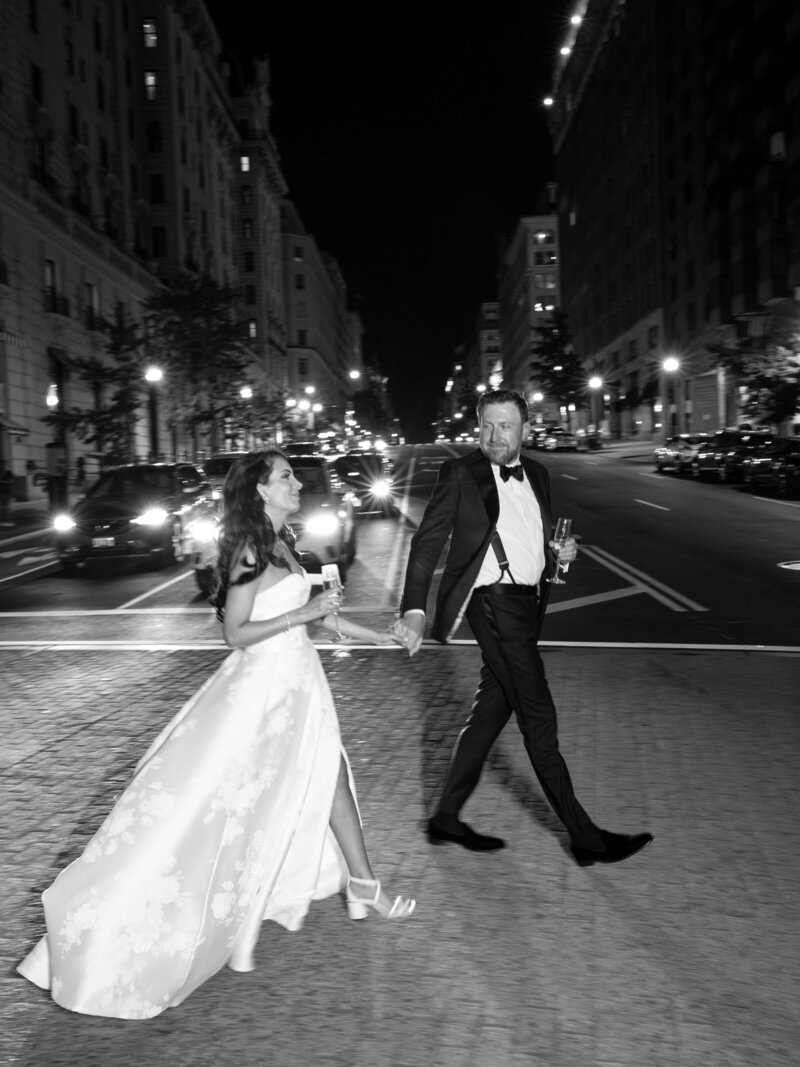 A couple dressed in formal attire walks hand-in-hand across a city street at night. The woman wears a long gown and heels, and the man wears a tuxedo. Both hold drinks, smiling as cars and buildings light up the background. Black and white photo.