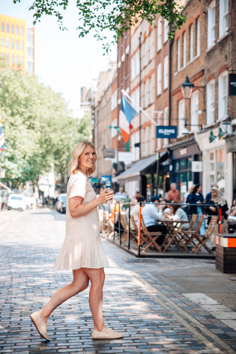 Emily Collins smiling while walking on a cobblestone street with an iced coffee, wearing a light dress and casual shoes