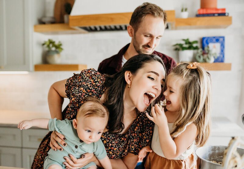 mom holds baby and leans over to take a bit of food her young daughter has, dad stands behind them