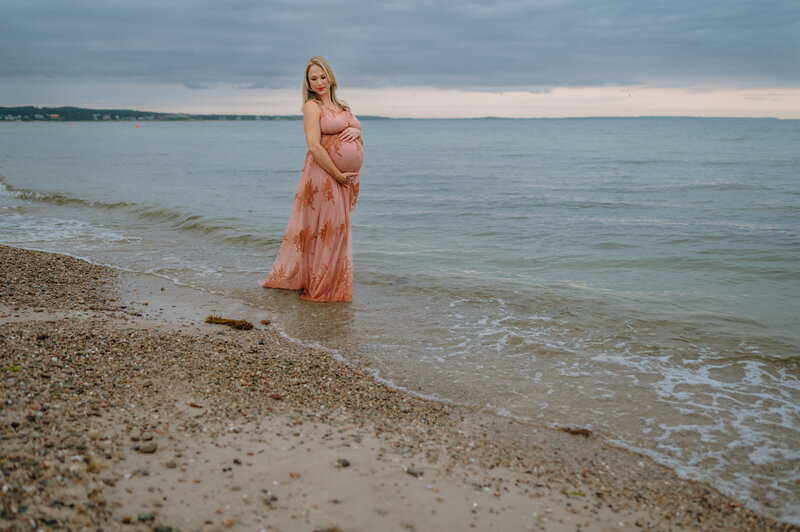 Pregnant mother on a beach holding her belly in a pink maxi dress.