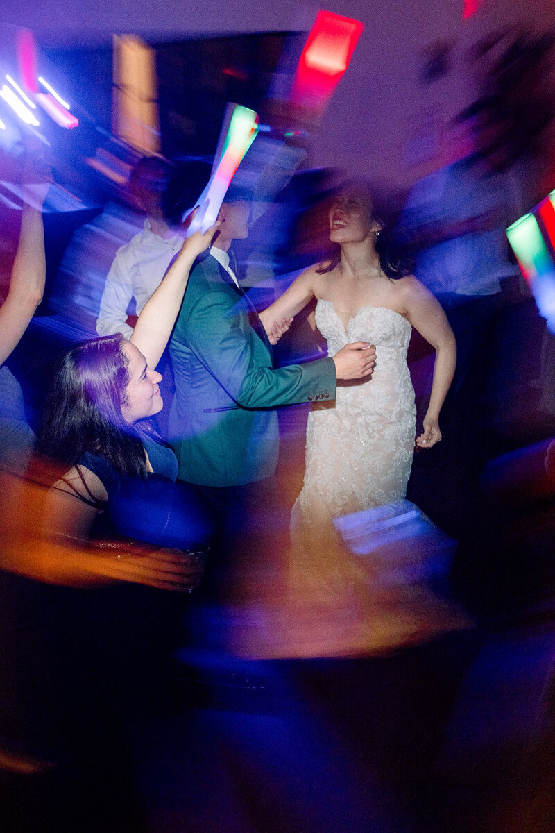 The couple shares a joyful moment on the dance floor at their California wedding reception. Surrounded by friends and family, the room comes alive with vibrant lights and energy as the newlyweds dance together. The bride’s lace gown and the groom’s suit are beautifully illuminated, capturing the lively atmosphere of celebration. This dynamic image encapsulates the excitement and love felt during the couple’s special