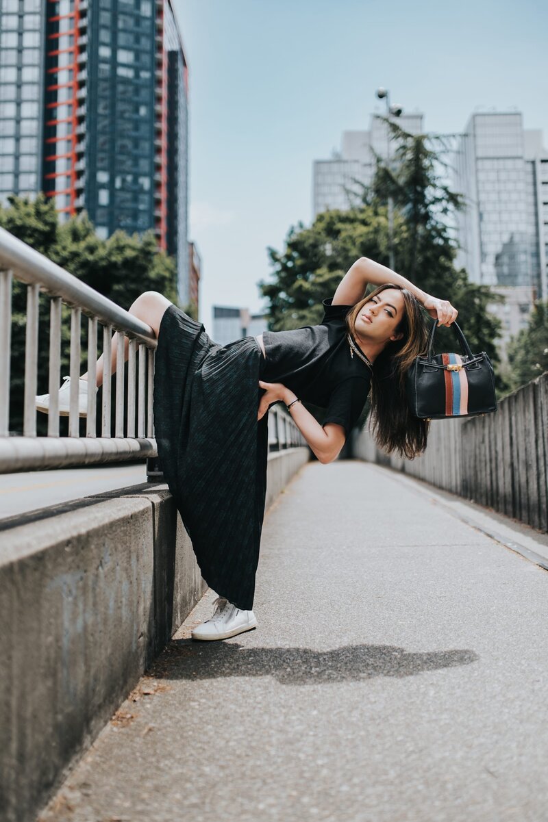 Woman wears a black, long dress and strikes an editorial pose holding a purse outdoors.