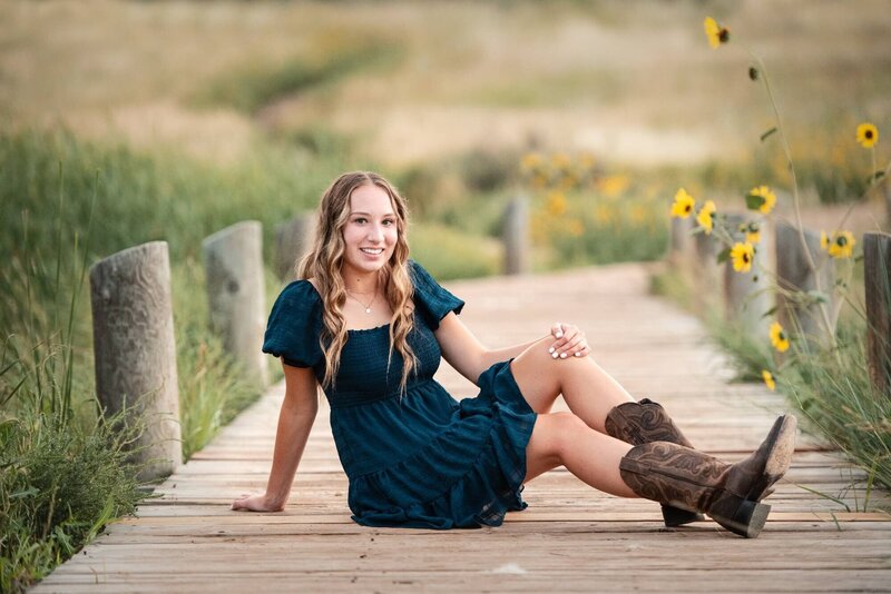 A high school senior in a blue dress sits across a wooden boardwalk at sunset with yellow wildflowers