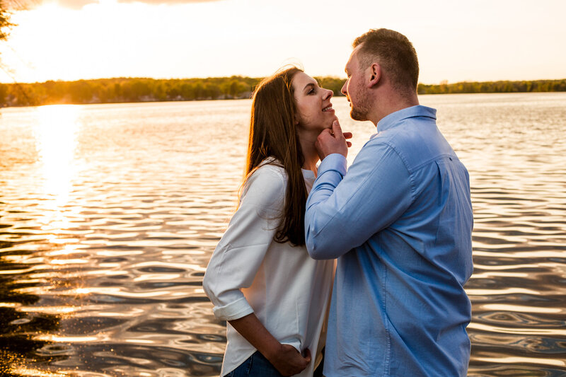 Man lifts fiancee's chin to give her a kiss next to Edinboro Lake near sunset