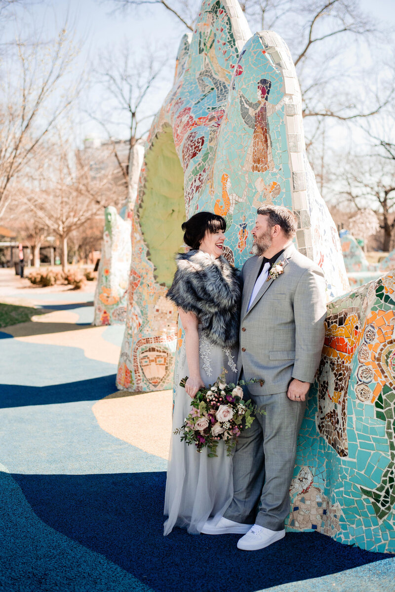 Bride and Groom standing near mosaic instillation, smiling at one another