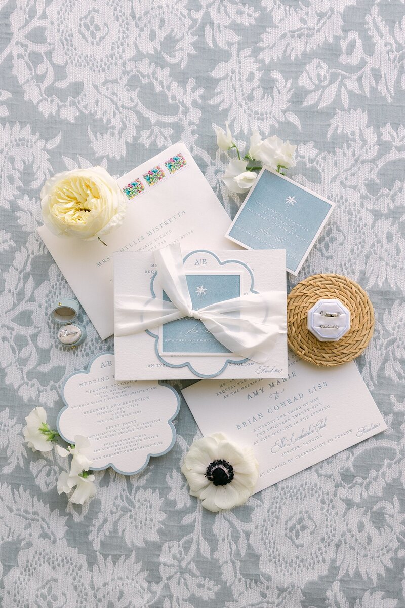 A flat lay featuring an invitation suite, set on a soft blue lace backdrop.
