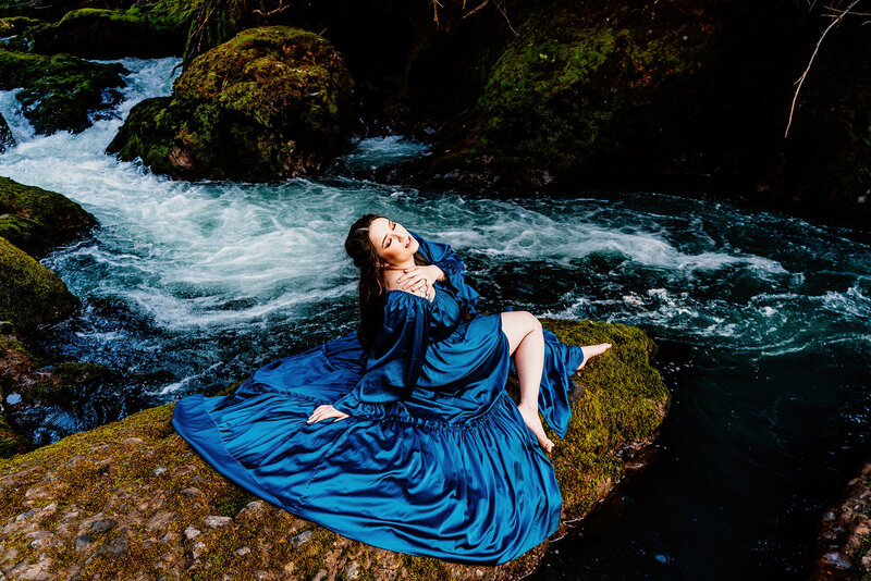 a model sitting on a mossy rock with a waterfall behind her