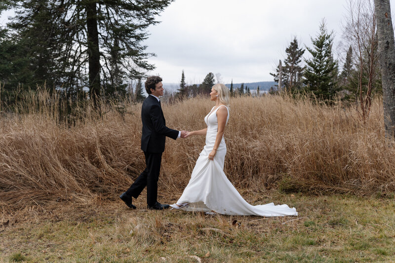 Couple eloping in nature walks toward each other, reaching out gently, framed by water and tall pine trees. 