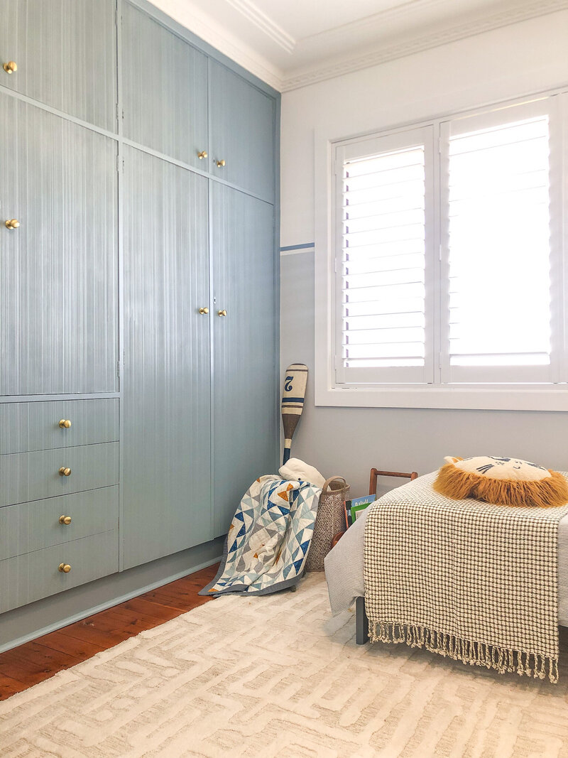 Cozy room featuring custom interior design in Newcastle, with blue cabinets, a patterned rug, and pillows on a bed next to a window with shutters.
