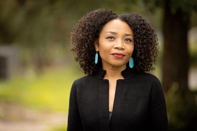 A female author with curly hair, wearing turquoise earrings and a black top, stands outdoors. She looks slightly upward with a calm expression—a style often captured by a skilled Jacksonville headshot photographer for business headshots in natural settings.