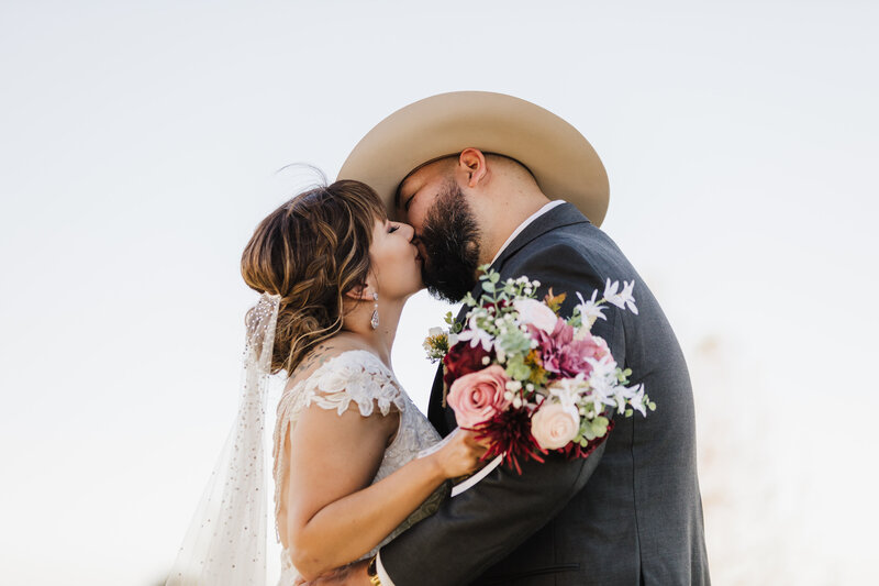 Man and woman kiss each other at their wedding while the woman holds a bouquet of flowers