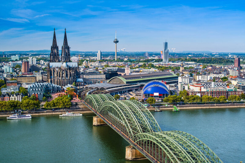 A cityscape featuring a large Gothic cathedral with twin spires, a green arched railway bridge crossing a river, and a sprawling urban area under a clear blue sky.