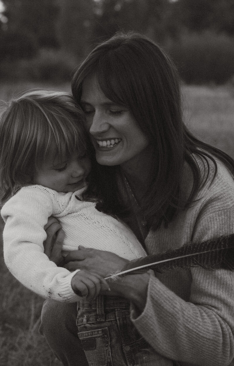 A husband and wife holding hands with their 2 daughters and son, smiling and walking towards the camera. They are outside with trees around them during Fall.