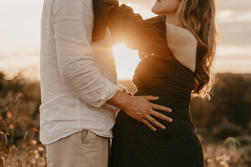 Pregnant woman in a sheer white dress posing on a sandy beach during a sunset maternity session in Old Bridge, NJ, captured by Burkat Photography.