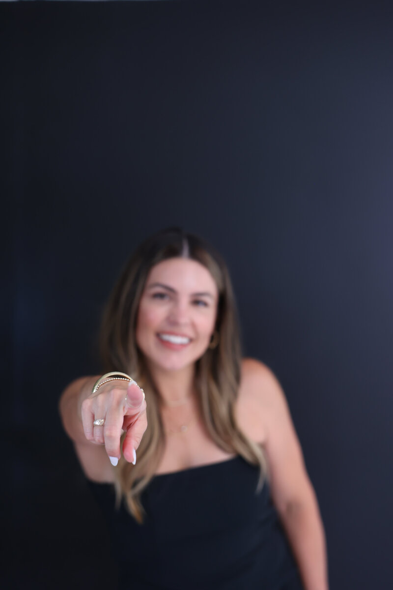Smiling woman in black outfit pointing directly at the camera against a dark green background, encouraging personal empowerment.