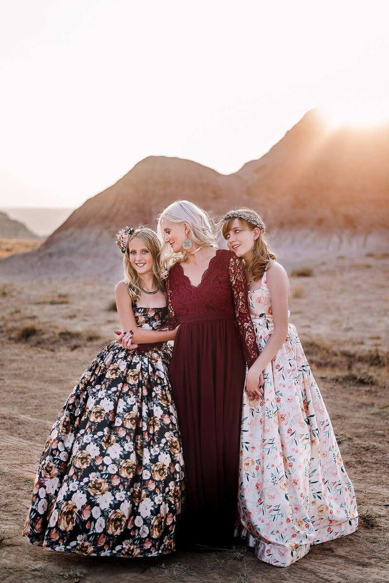 Blonde mom wearing a red gown smiles at her daughter while holding hands with her other daughter in the red rock canyon in Wyoming