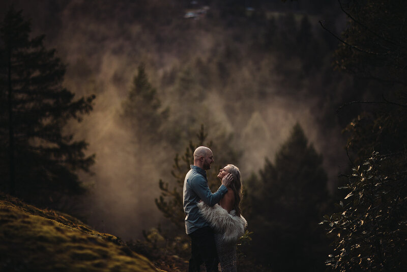 A white couple face each other and look into one another's eyes. His hand is on the side of her face. The background is a lush green forest with a lingering fog.