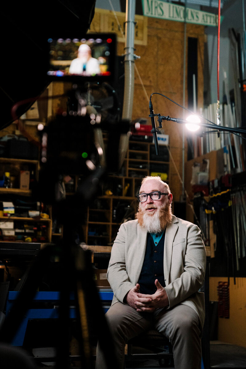 a man in a red and grey beard sits under a boom microphone in front of a camera and display