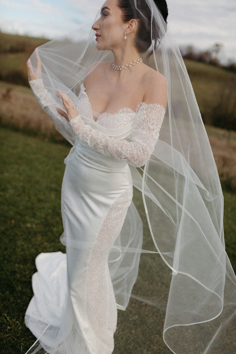 A woman in a white lace wedding dress and veil stands on grass, hands adjusting the veil at a Farmhill wedding in Peterborough. Adorned with a pearl necklace, she is captured beautifully through wedding film photography, set against a blurred landscape captured by Toronto wedding photographer Jennifer van Son.