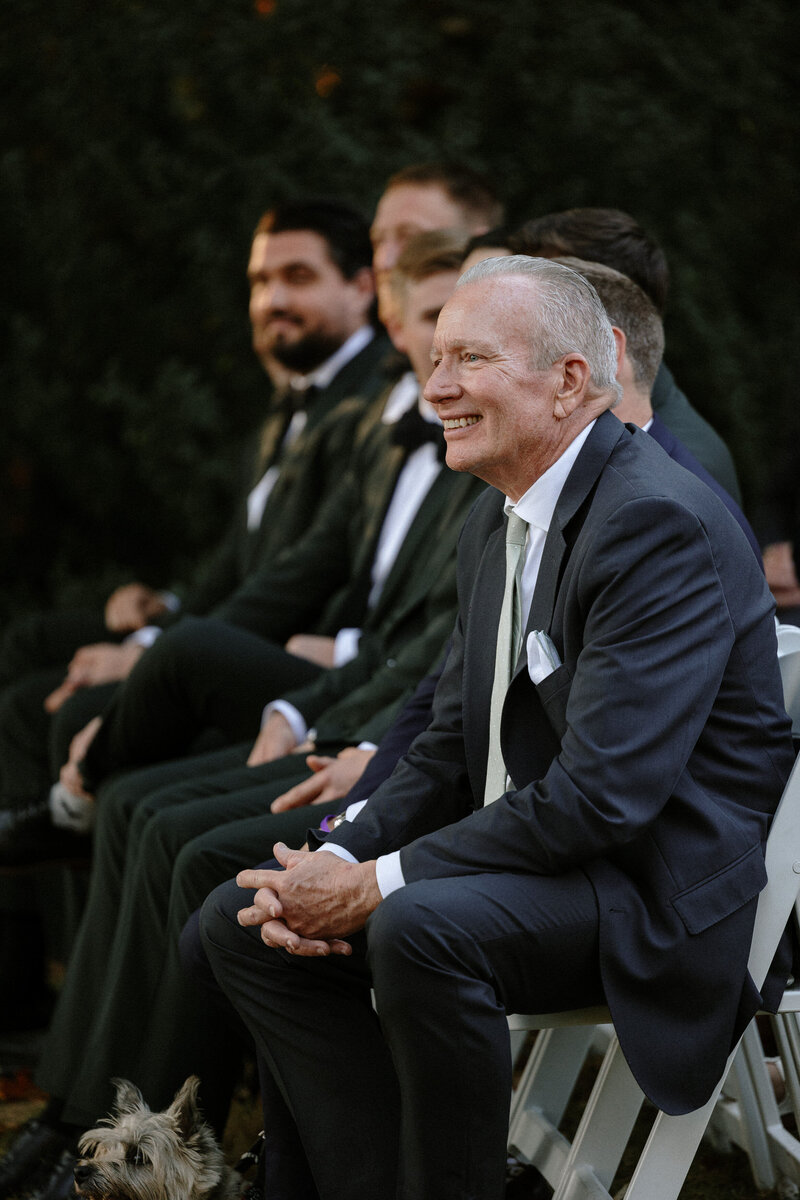 A group of men in formal attire seated outdoors, with a small dog resting at the feet of one man.