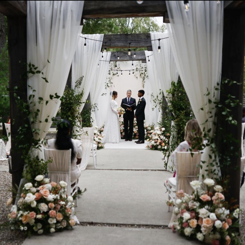 An intimate indoor wedding ceremony in a stone-walled room with a vaulted arch. The couple stands in front of a wooden mantel adorned with candles, surrounded by seated guests. Tables with floral arrangements are visible in the foreground.