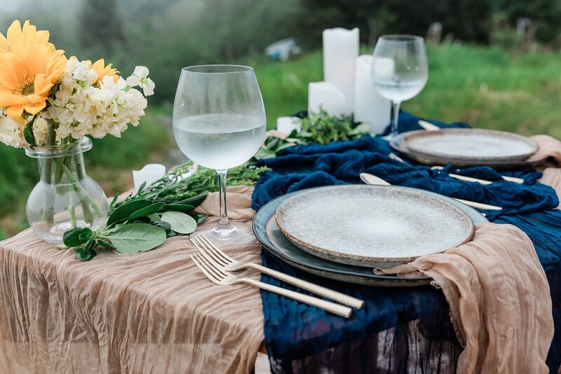 Two glasses of frosty glacier water sit in sparkling wine glasses--they are details from a luxury helicopter and glamping elopement package at the deck of a luxury glamping tent in Alaska.  On the small decorated sweetheart table is a light tan and deep navy blue gauze table linens, earthen pottery inspired plate sets, and glowing gold silverware.  with matching gauzy tan napkins.  There are white candles, and a hit of yellow flowers visible on the edge of the table, with lush green grasses visible off the edge of the yurt's deck. 