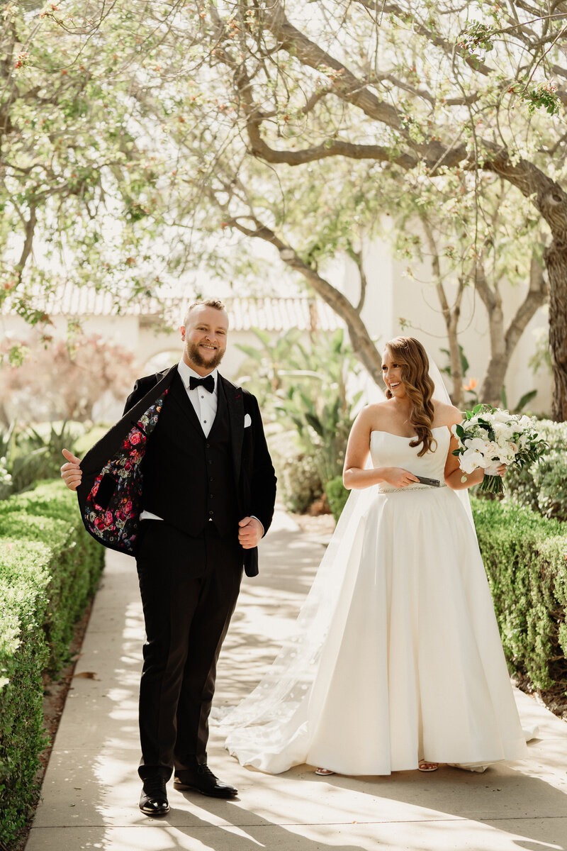 bride wearing a white dress and a cathedral polka dot veil holding a large all white bouquet and groom wearing a custom black velvet wedding tuxedo showing off the custom lining on the inside of his jacket at spanish hills country club captured by los angeles wedding photographer magnolia west photography