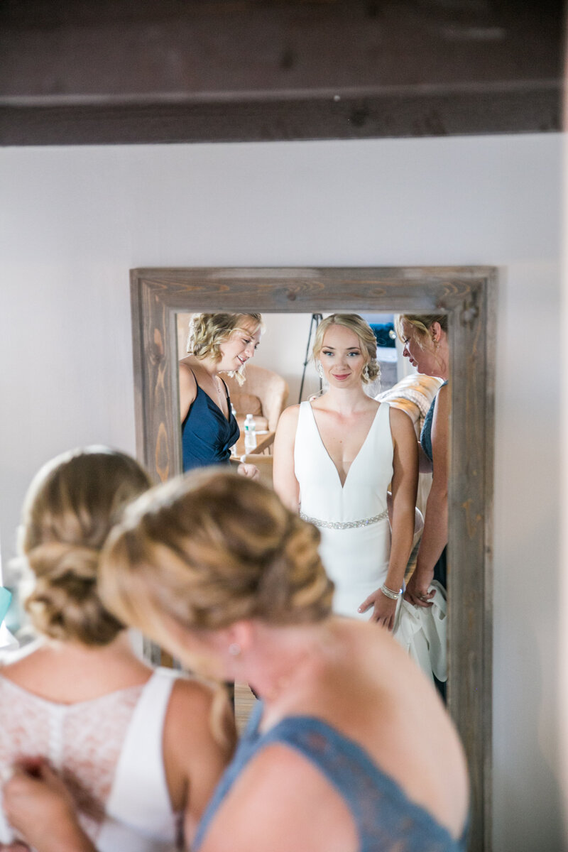 Photo of Bride looking in Mirror with Bridesmaids helping with dress | Grace on the Lake Wedding Photography