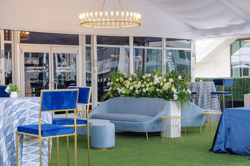 Lounge setup with blue furniture, floral arrangements, and a chandelier under a tent.
