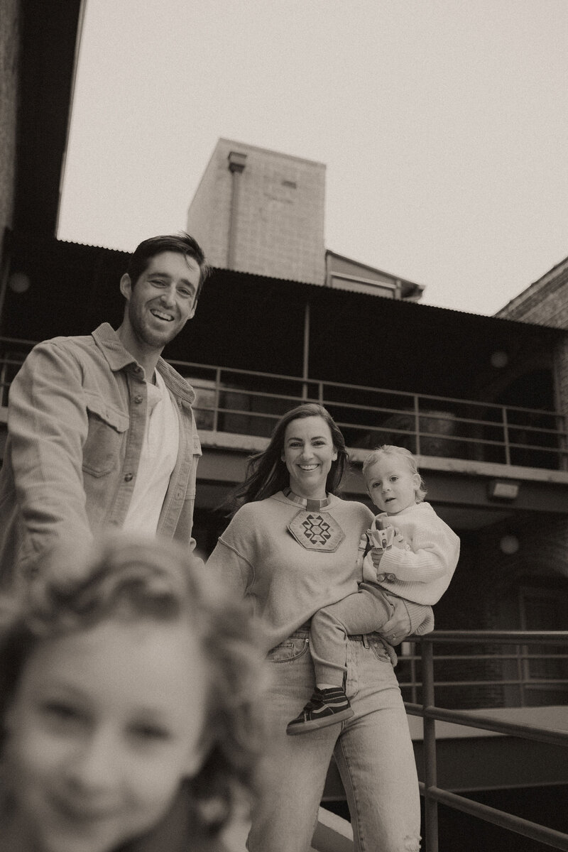A father and mother holding her baby run after their toddler who is racing away from them. 