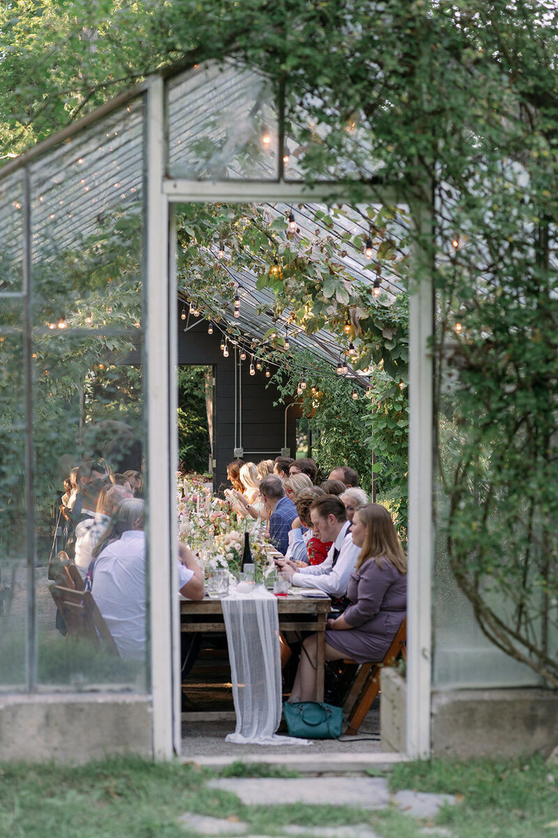 A candid peek inside the Glasshouse Community greenhouse showing guests enjoying an intimate summer wedding dinner surrounded by lush florals and greenery.