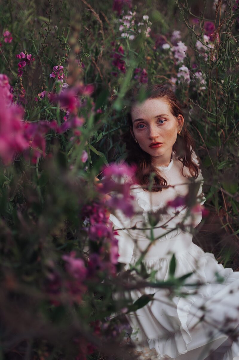 Young, red-headed woman sits in a flowy white dress in a field of flowers 
