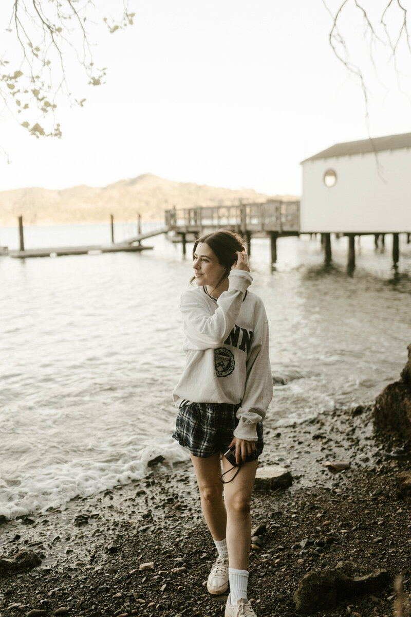 Woman wearing a vintage crewneck, skirt, and white crew socks with tennis shoes standing on beach in front of ocean dock