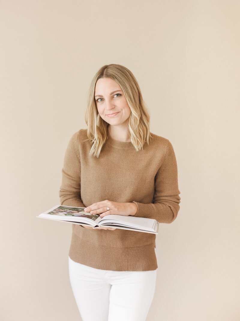 A woman sitting standing in front of a concrete wall smiling and looking into the camera.