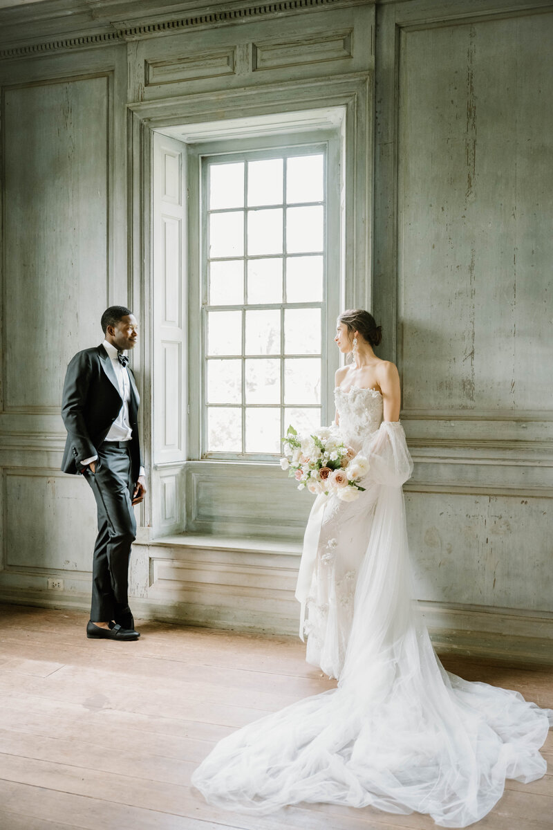 Wedding Photographer RI | Bride in a flowing gown and groom in a suit share a tender moment by a large, sunlit window in an elegant, vintage room.
