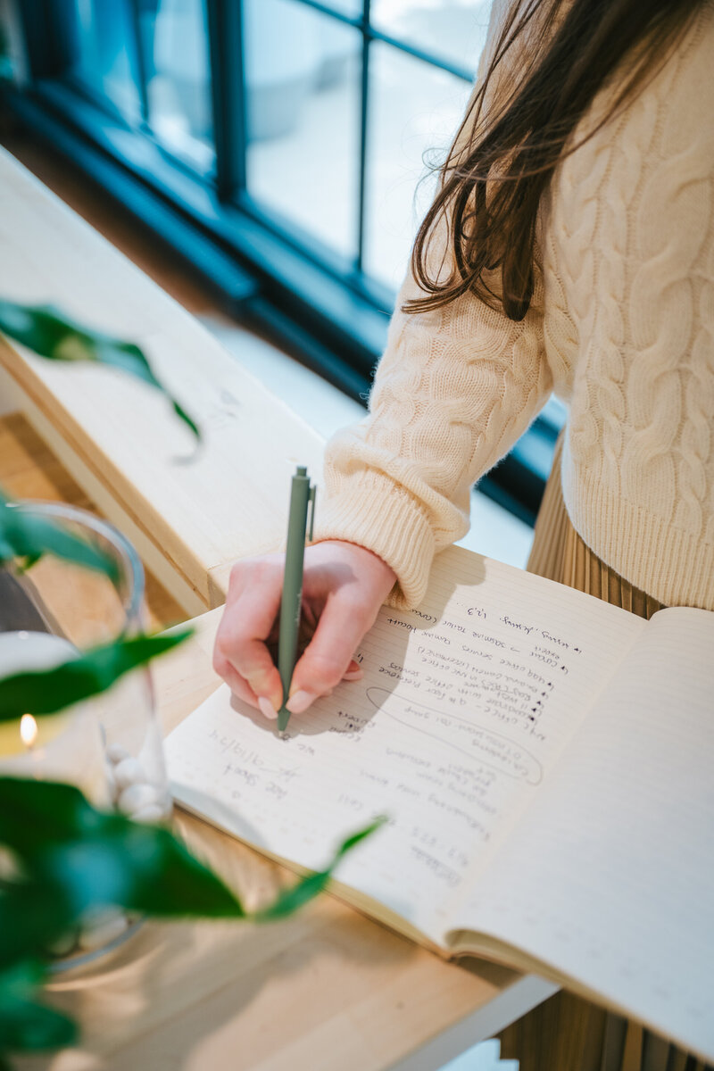 Woman in cream sweater writing in a notebook with a green pen