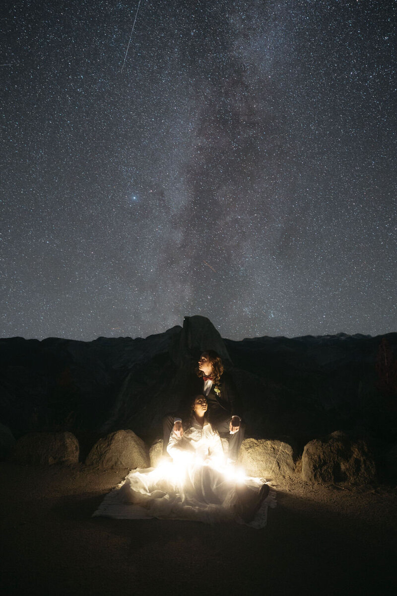 Unique Elopement Ideas | Eloping couple sits on a rock together illuminated by string lights with the milky way stars in the sky and Glacier Point behind them