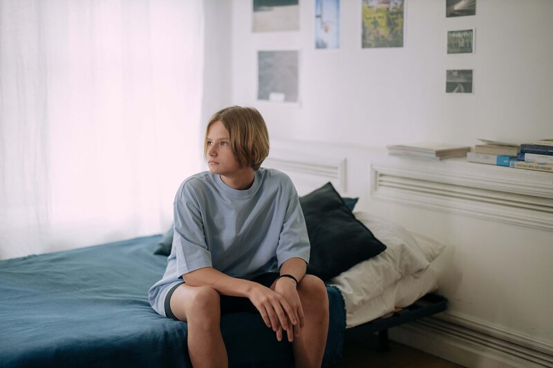 A teenager sits on their bed, arms resting in their lap. They look blankly past the camera, over their right shoulder.