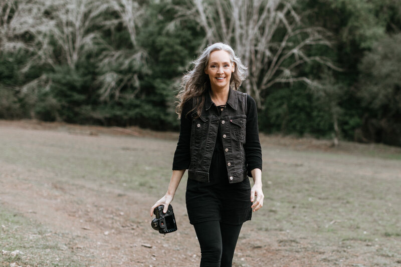 portrait of a Marin family photographer smiling and holding camera in a bright white room. She is wearing a black vest and black shirt and has silver hair, she is leaning toward the camera.