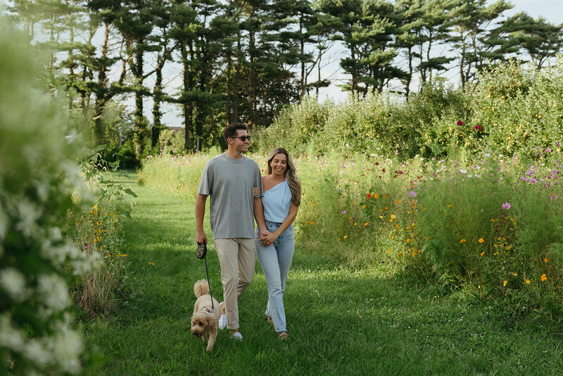 couple walking with their dog in grass during engagement photos, captured by Elsie Goodman, an NYC engagement and couples photographer