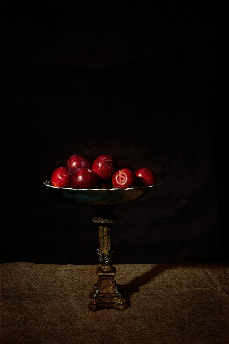 A moody still life featuring red plums elegantly displayed in an antique bowl, set against a dark background. Captured by Mica McCook, this image evokes classic still-life paintings with its rich shadows and dramatic lighting.