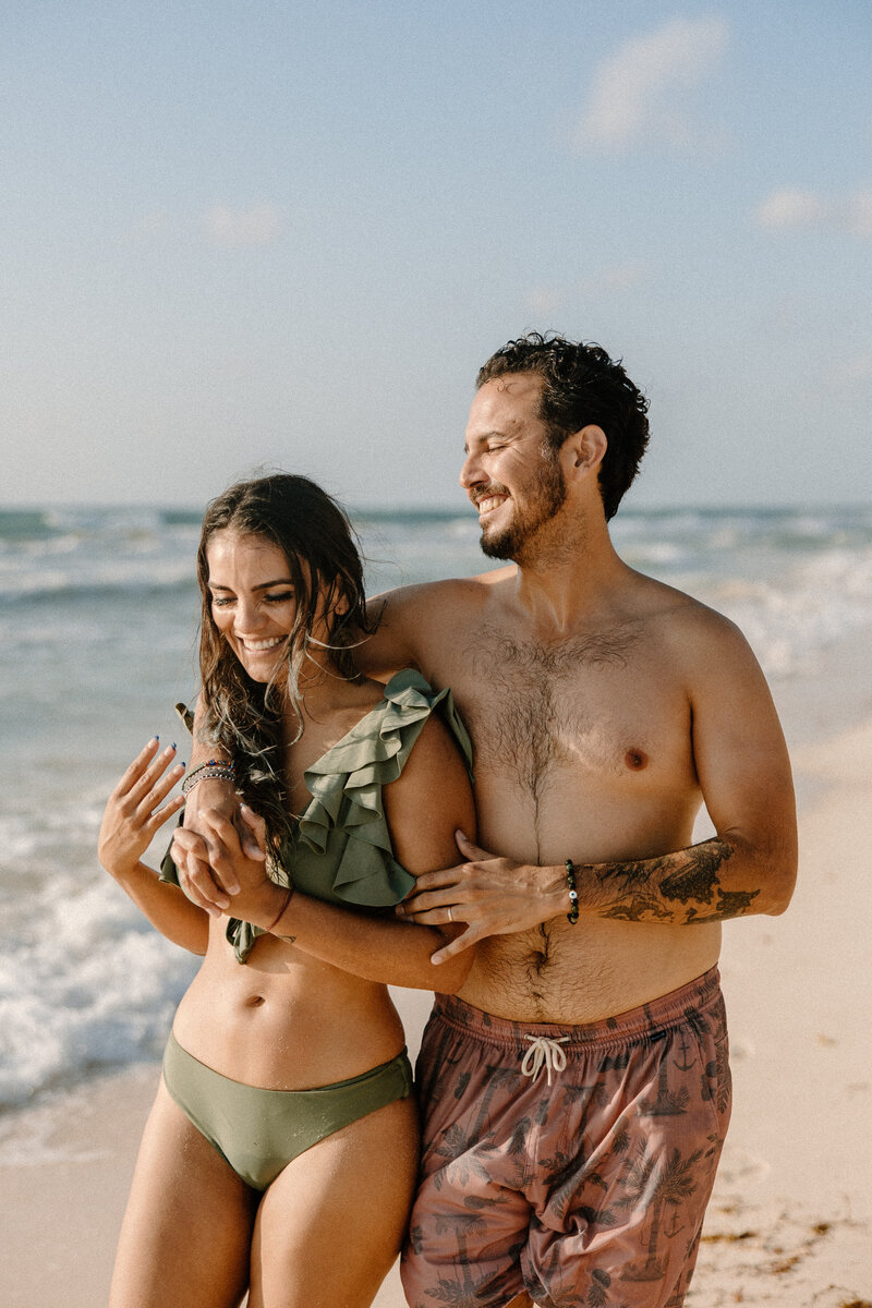 Couple laughing and embracing on the shoreline during their Tulum engagement photoshoot, waves crashing behind them.