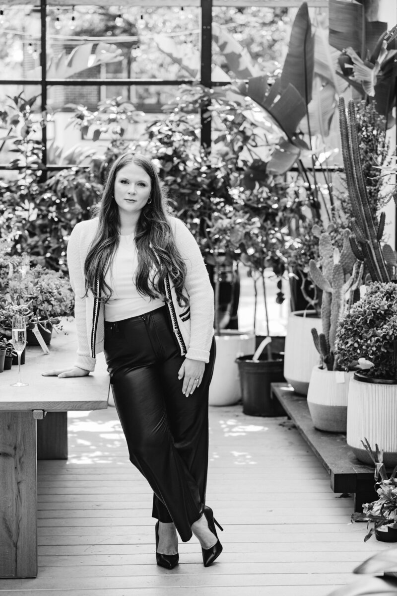 Black and white photo of virtual assistant agency owner Kate standing in a room pull of plants leaning against a table