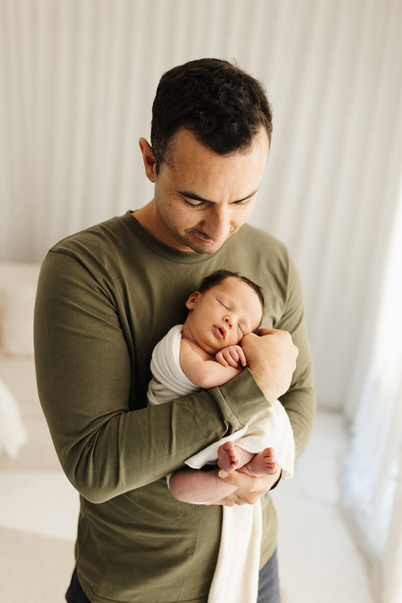 A father wearing a green long sleeve top holding his newborn baby while photographed by Christchurch photographer Lucy Jane Photography