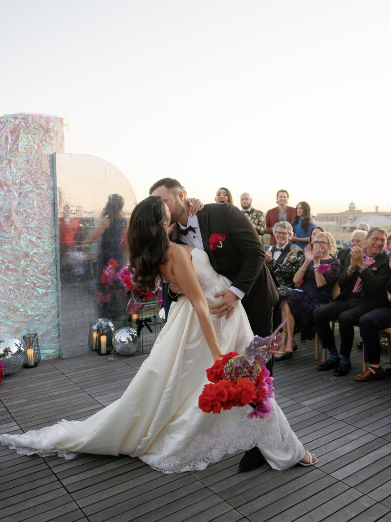 bride and groom kissing at the ceremony on the terrace in dc in front of an iridescent wall backdrop surrounded by couples loved ones