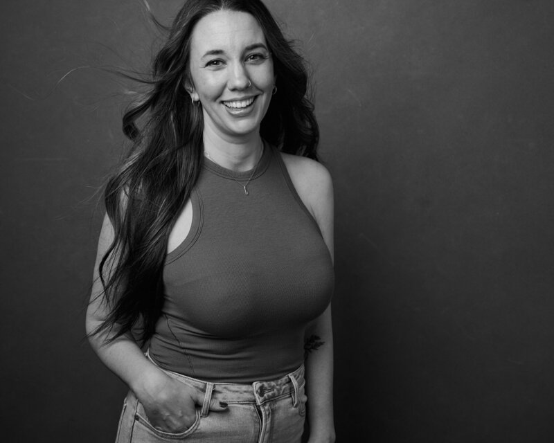 Black and white portrait of a woman with long, flowing hair, smiling brightly as strands of hair catch the air. She wears a fitted sleeveless top and high-waisted jeans, her hand casually tucked into her pocket. A radiant, unfiltered moment from 'THIS IS ME – A Photography Experience by Katie Campbell' in Atlanta, GA, celebrating confidence and authenticity.