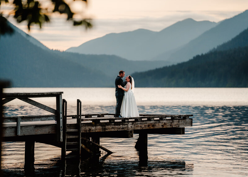 a couple in their wedding attire stand at the edge of a dock. The lake shimmers below them and mountains roll behind them illuminated by the sunset. 