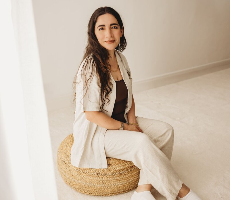 Woman with long dark hair, wearing a beige linen outfit, sits on a woven stool, looking at the camera.